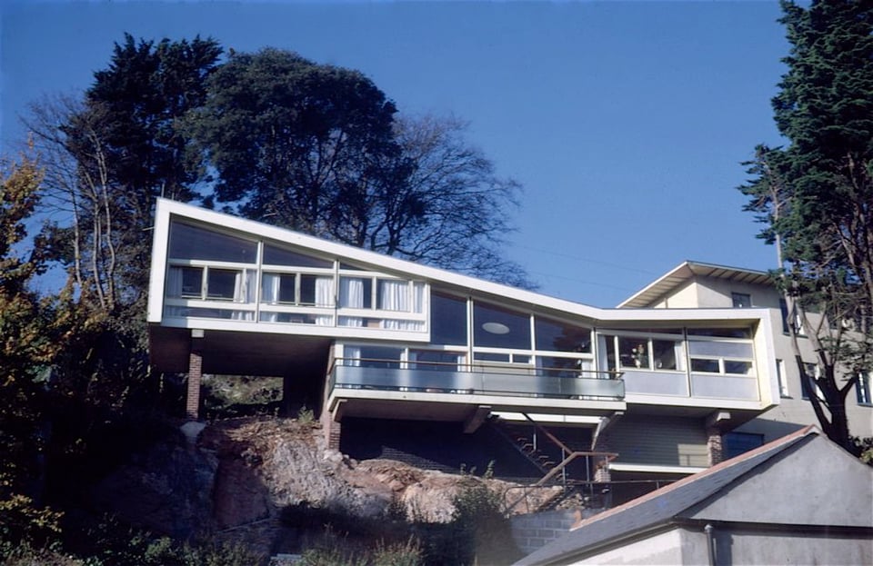 A midcentury house, built into the side of a cliff edge within a town. It has an asymmetrical roof in a V shape. Beneath that are split level rooms and a projecting balcony. Steps lead down from the balcony across the face of the rock.