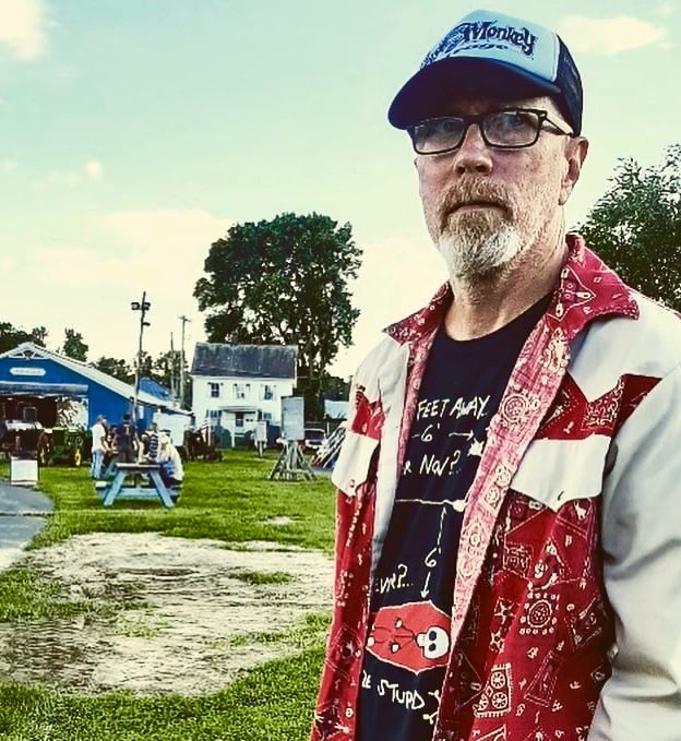A man with glasses and a beard wears a trucker cap, a snap-front western shirt and a black t-shirt.