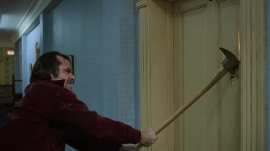 Jack Torrance attacking a beige-coloured door with an axe, gritting his teeth in a look of rage.