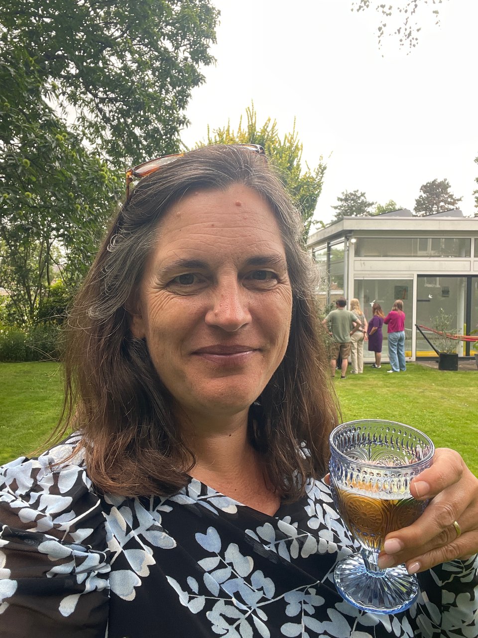 A closeup of a woman smiling at the camera holding up a glass of PimmsCup as she stands on a lawn in the garden.