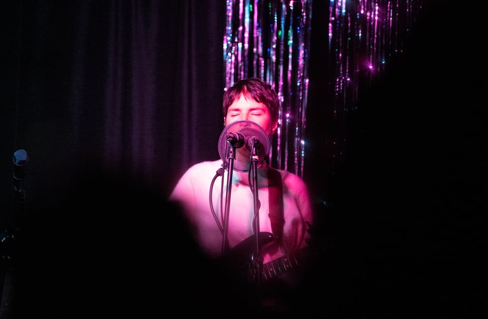 Photo taken before the performance that Alive People by Ruth Garbus was recorded at. The image features Ruth Garbus singing with eyes closed and a guitar, with a pop filter and two microphones in front of the lower part of her face, and sparkly stage curtains behind her.