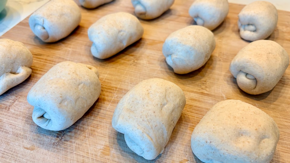 Three rows of four rolls of dough on a wooden board, ready to be turned into bagels