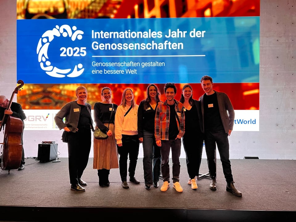 A photo of a group of people standing on a stage, posing for the camera, with a projection in the background that says “2025: Internationales Jahr der Genossenschaften”