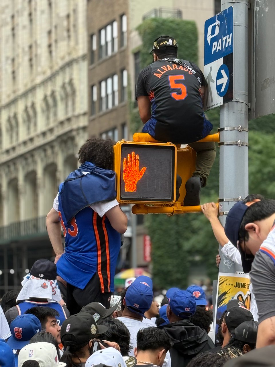 photo of Latino man in Jose Alvarado Knicks jersey, sitting atop a walk/don't walk traffic light. A man in a Patrick Ewing jersey is below, and they are flocked by many thousands of Knicks fans