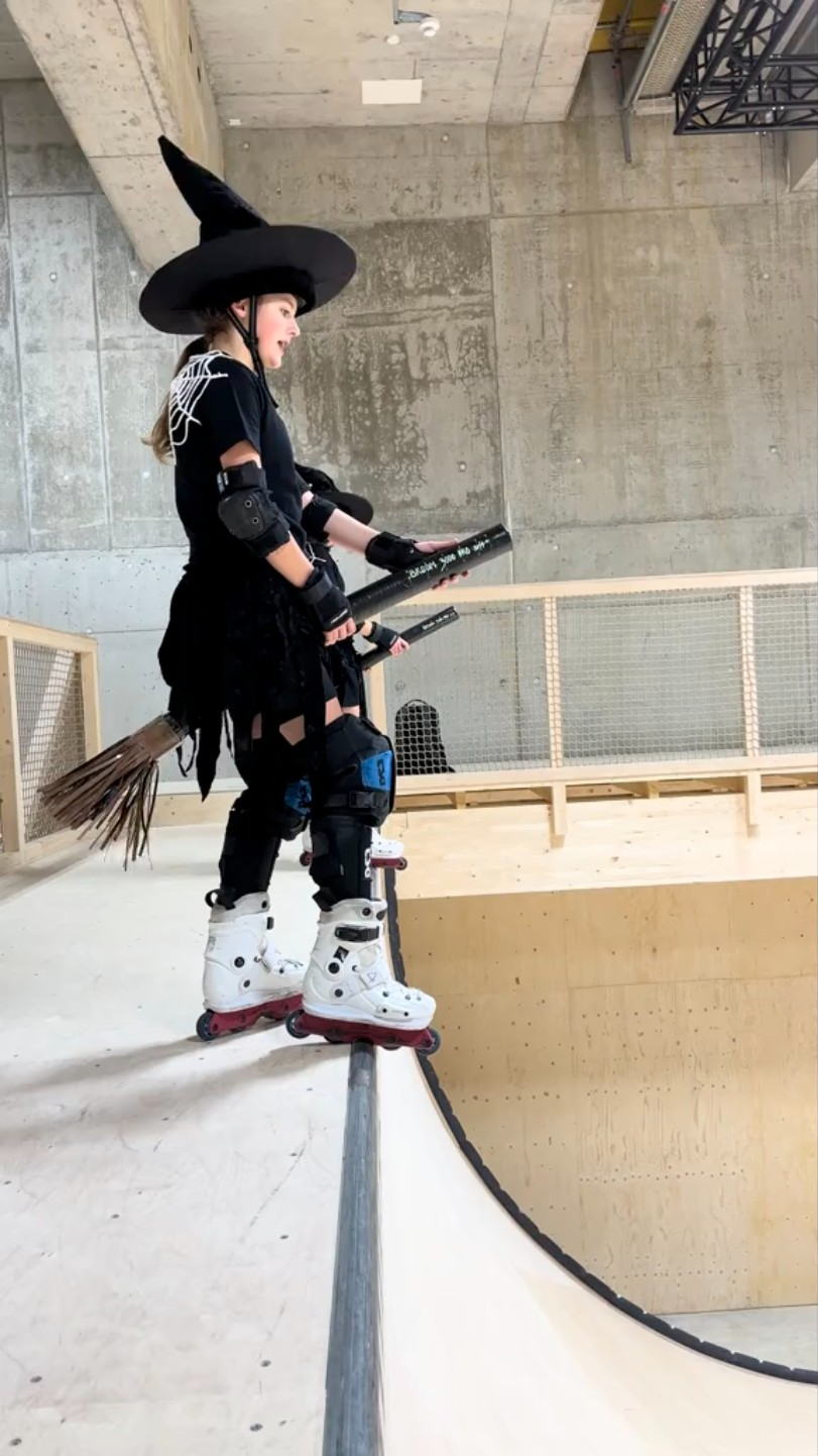 Two young women stand at the top of a skating half-pipe. They're dressed as witches and holding brooms