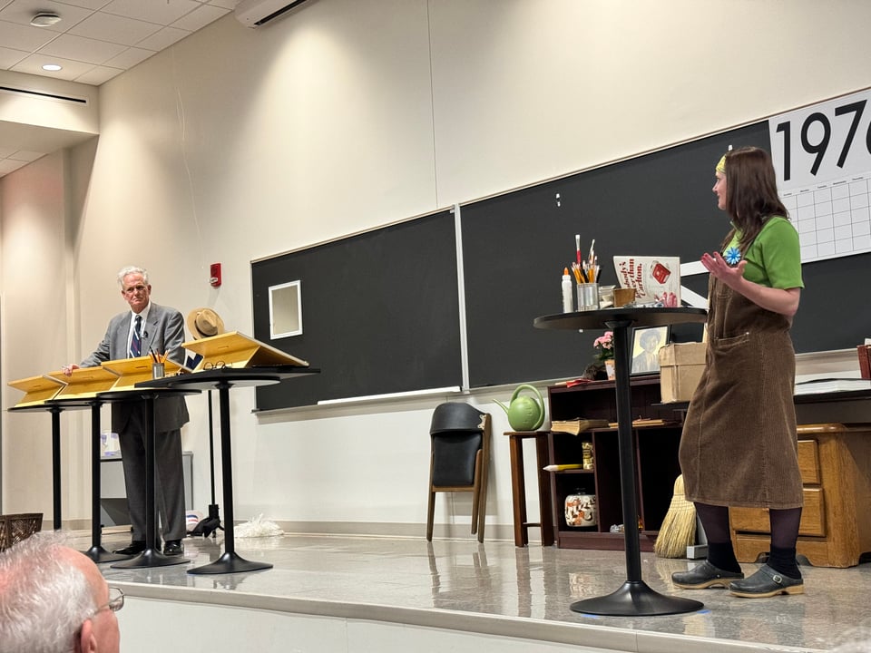 Photo of Tom Gammill and Caitlin McGurk performing in “A Morning with Ernie Bushmiller”: picture shows shallow stage with Gammill at far left dressed as Bushmiller in front of several drawing boards; McGurk far right in period dress in front of a sign reading 1976