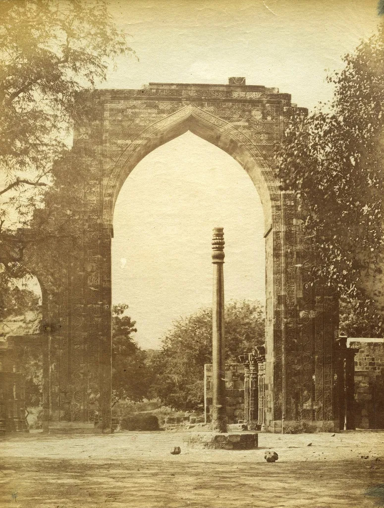 ChatGPT said:A sepia-toned historical photograph shows an ornate stone archway framing a tall iron pillar in the center of an ancient courtyard. The arch is richly carved with intricate geometric and floral patterns, typical of early Indo-Islamic architecture. Surrounding trees and scattered stones suggest the site is part of a centuries-old ruin