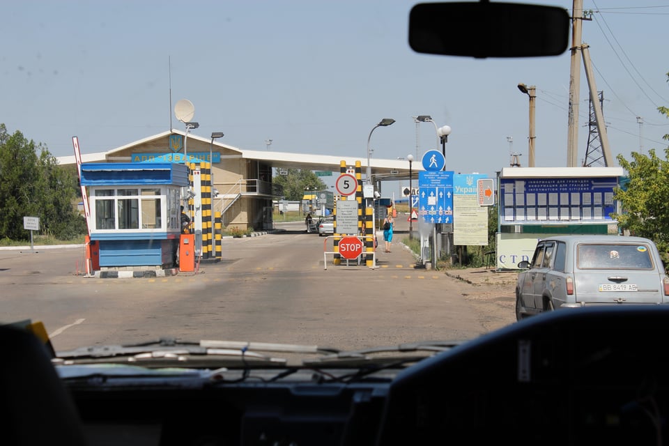 Photograph taken through a vehicle windscreen of a small border checkpoint with Stop sign, guard post and vehicle inspection points.