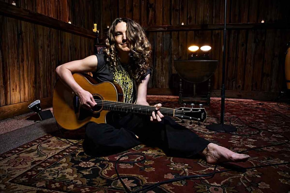 A woman with curly hair cascading past her shoulders sits on an oriental rug in a dimly lit room, holding an acoustic guitar with her left leg outstretched.