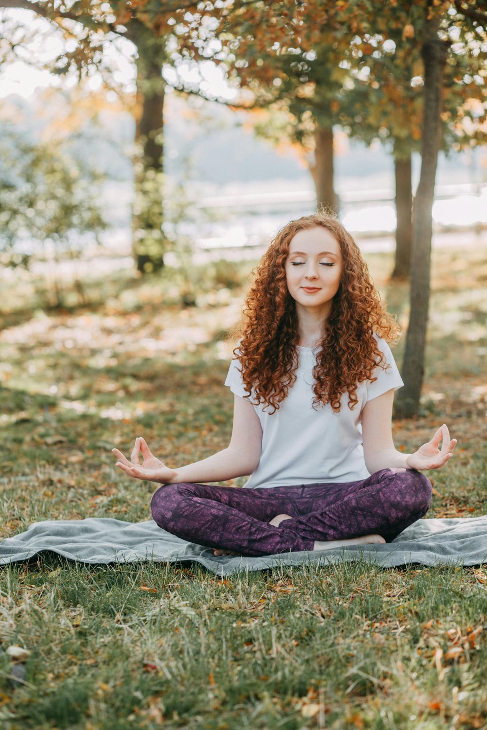 A young, white, redheaded woman in a white shirt and purple yoga pants meditates on a towel outside.