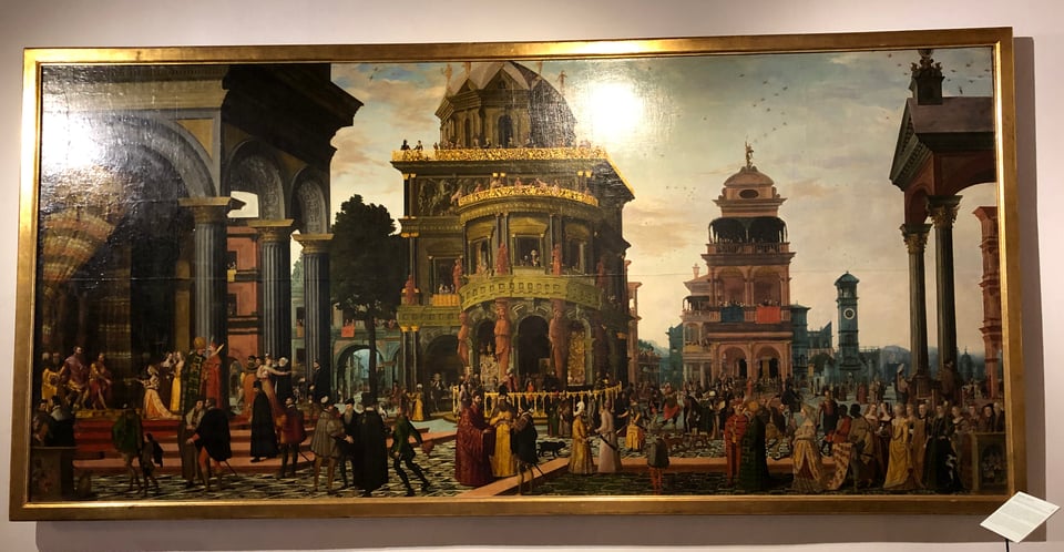 A framed painting on a wall. It depicts landscape-shaped scene of a sixteenth-century European city. Well-dressed people mill around several monumental buildings with arches and columns.