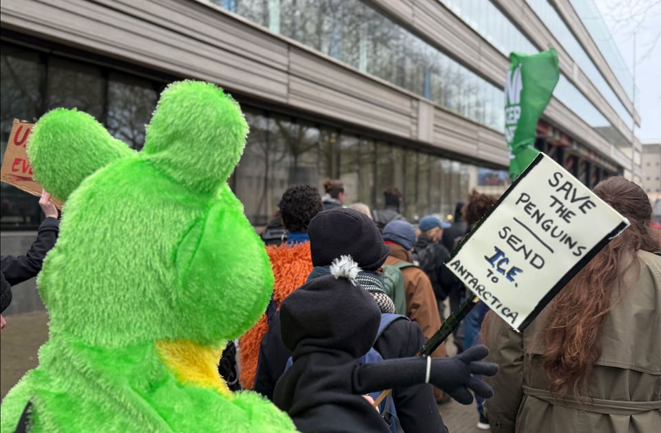 A person in a frog suit holds up a small sign that says "Save the Penguins - send ICE to Antarctica"