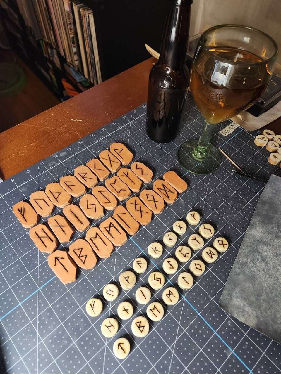 A cutting mat on a work table, and on the mat are arranged two sets of Elder Futhark runes, one with leather tiles and the other from a slim wooden branch.