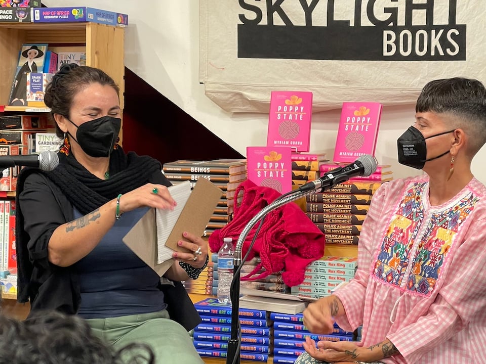Olga García Echeverría and Myriam Gurba seated at Skylight Books, both masked, in conversation. Olga is holding up some cards that contain questions and Myriam is about to choose a card. They are seated in front of many stacks of books.