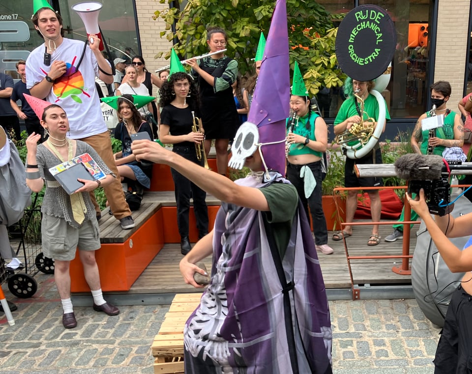Someone dressed in a skull mask and purple robe holds an iPhone aloft while grasping a rock with the other hand. In the background is a crowd of musicians and onlookers.
