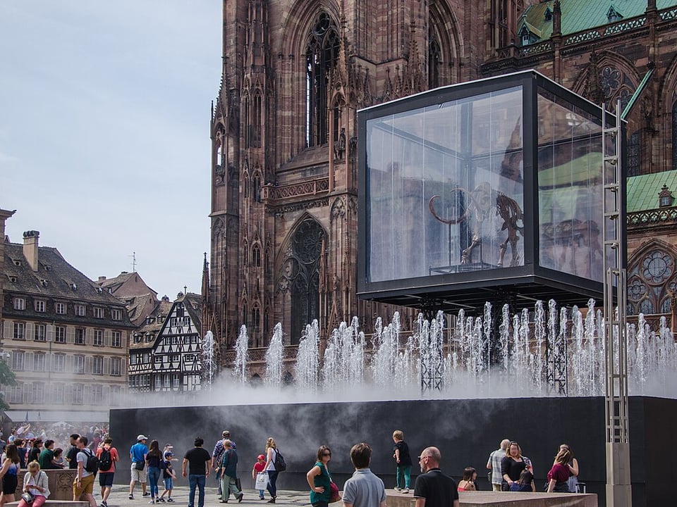 A group of people milling about a plaza in front of what appears to be a large cathedral. There is a water fountain display with dozens of water jets shooting straight up, and a glass cube raised into the air above them. Within the cube there is the skeleton of a creature with large tusks, perhaps a mastodon of some kind. It appears as if it is overlooking the crowd.