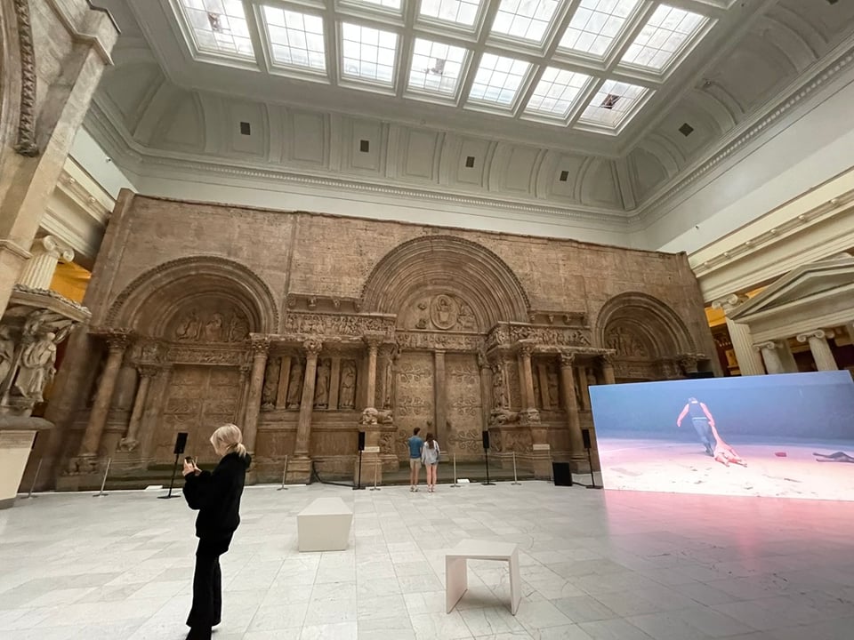 a woman photographing something to the left, standing in front of the sculpted facade of a church, inside a museum gallery