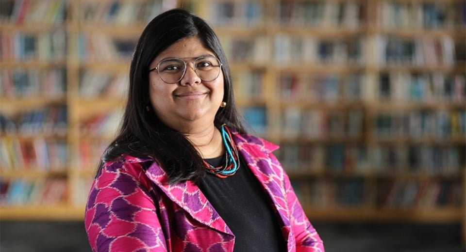 JMC Prof. Sejuti Das Gupta photographed in the JMC Library wearing a black shirt and pink/purple jacket.