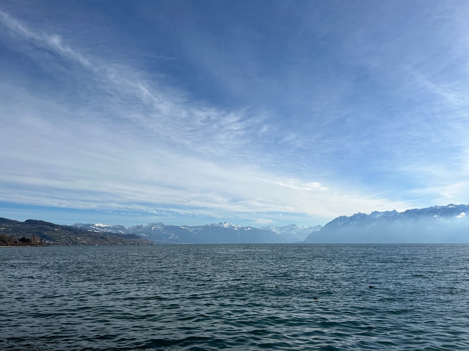 A photo of a lake, with snow capped mountains in the background. There are wispy clouds in the sky, and the photo is overall very blue.