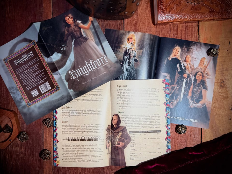 Three copies of the Knightcore zine on wood, two open, amid dice, a book, a metal helmet, and velvet.