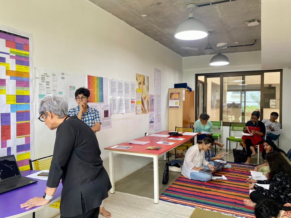 A group of workshop participants, some seated on the floor, others in their chairs, and still others standing and engagement with the workshop materials on display.