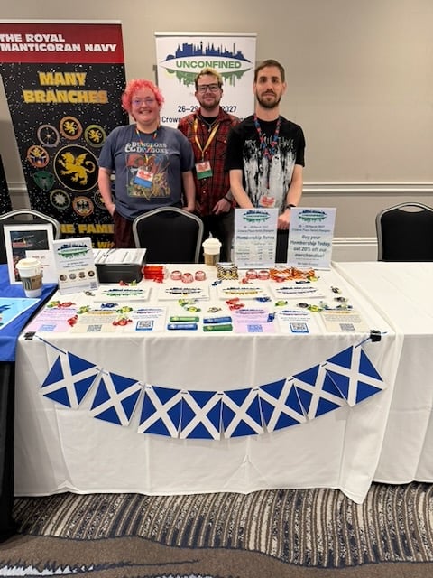 Three people standing in front of a table covered in leaflets, ribbons and sweets, with a ribbon of scots flags hanging down at the front of the table.