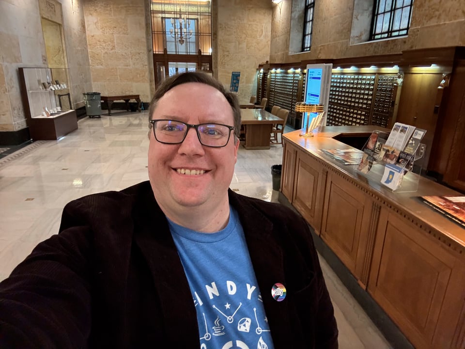 Middle aged white dude wearing a brown corduroy jacket over a blue Indy Hackers t-shirt. He has a Pride Ally pin on his left lapel and is taking a selfie in a lobby of the Indiana State Library.