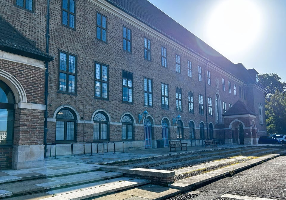 A large plain brick building with three floors and a heavy slate roof. The windows are square, like a cartoon drawing of a window. There are two porches at either end, which are square with a pyramidal roof.