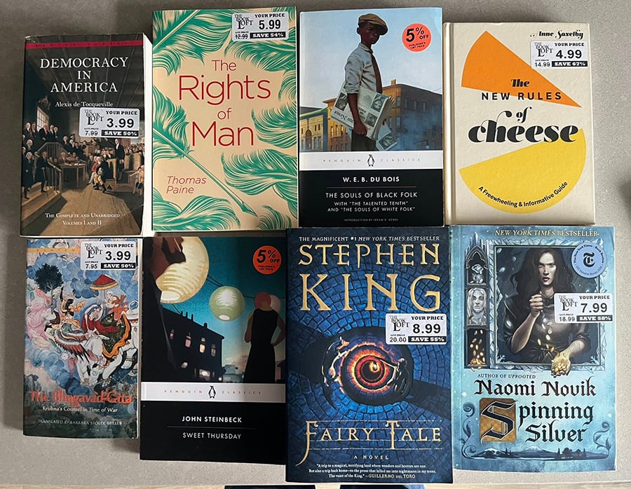 Eight books on a table: Democracy in America, the Rights of Man, The Souls of Black Folks, The New Rules of Cheese, The Bhagavad-Gita, Sweet Thursday, Fairy Tale, and Spinning Silver.