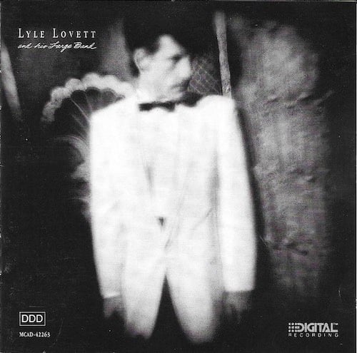A blurry black and white photo of Lyle Lovett, a white man with a long face and big floofy dark hair that dips over towards the front of his face. He's wearing a big white suit and dark tie that makes him look like a bandleader from the 20s or something. He looks to the right at something out of frame.