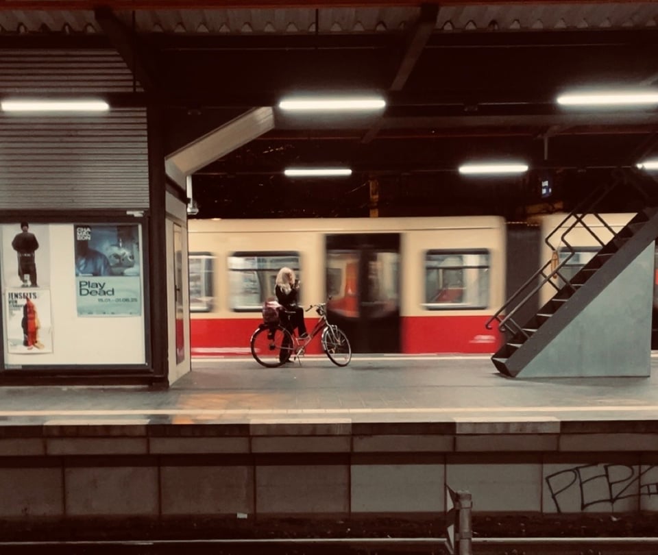 A cyclist with a black outfit and white hair, standing leaning against their bike in the middle of the frame on a train station. The train moves behind them, blurred. There is a half closed shop with advertisement on the left and a metal staircase on the right