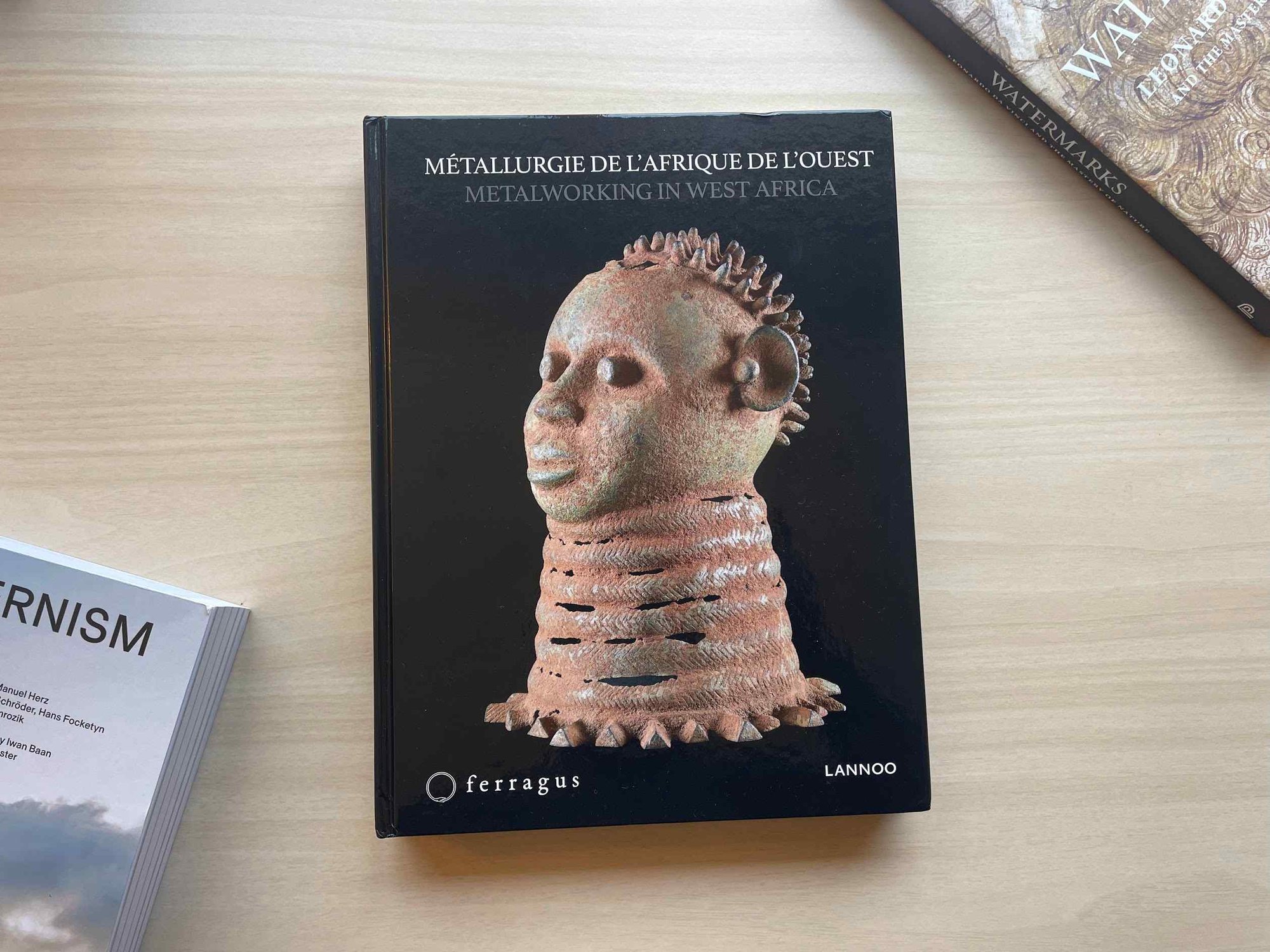 A hardcover book on a table, titled "Metalworking in West Africa" The cover art is a high resolution photo of a sculpture of a stylized human head against a dark background