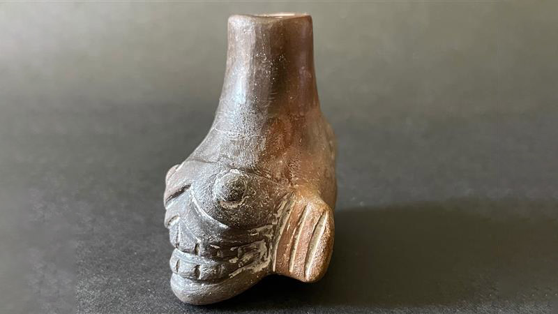 A picture of an Aztec, death whistle, which looks like a short, squat pipe, possibly carved from a large bone. the bottom of it has a the face of a monster card into it, with two large eyes and lips parted to show bared teeth