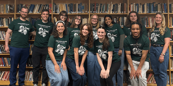 A group of JMC students and professors wearing matching green t-shirts pose for a photo in front of bookshelves. They are arranged in two rows, smiling and looking at the camera.