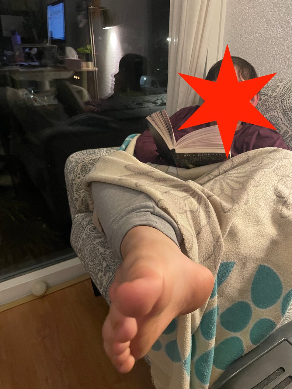 A boy sits in a comfy chair covered in a blanket reading a book. His bare foot is very close to the camera, so it's distortedly large.