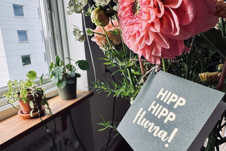 En bukett med blommor i köksfönstret. Ett kort är fäst i buketten med texten Hipp, hipp, hurra!