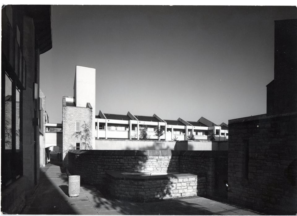 A two storey building with several flats along the top storey. The concrete frame has been built so the flats have a wide access balcony. They are brightly lit by the sun. Framing the view are the darker masses of the hall and the church tower.