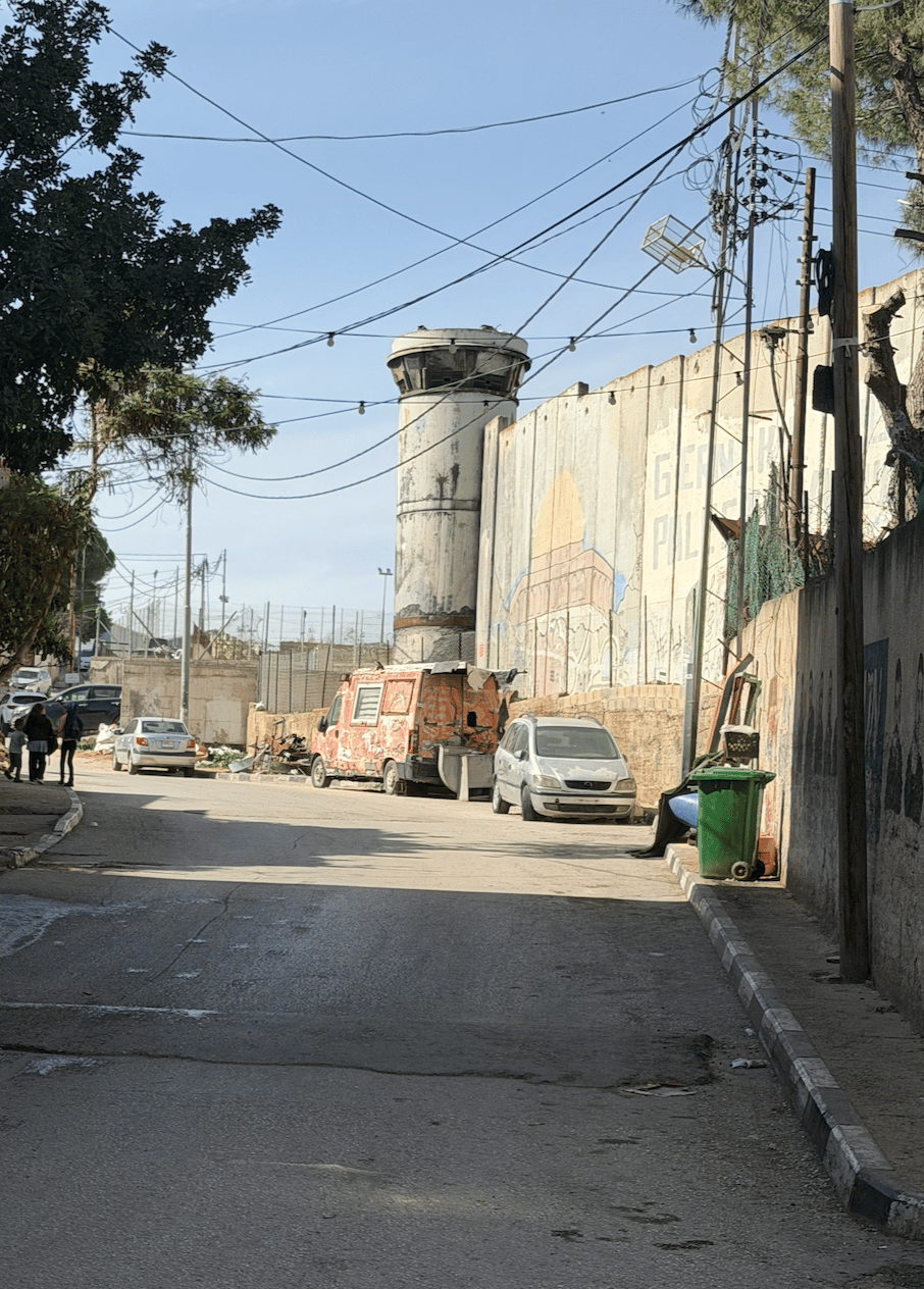 Photograph of a street featuring a tall concrete watchtower and a high concrete wall with faded graffiti. Several parked vehicles, including a rusted van and a small car, line the street, with overhead power lines and sparse trees visible under a clear sky.