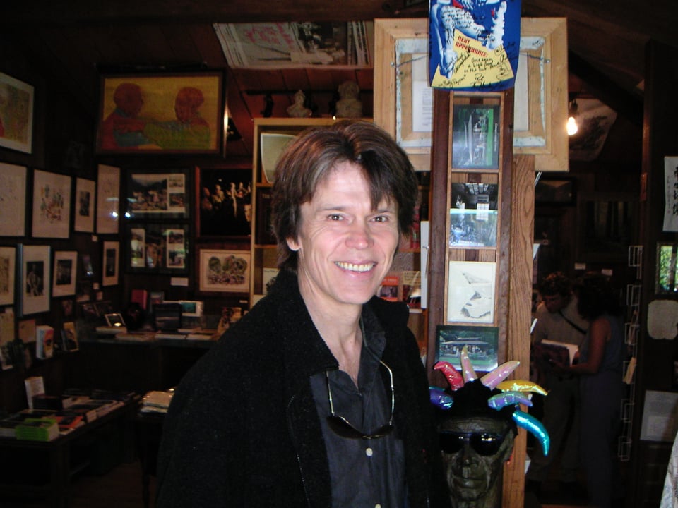 Photo of a while man with brown hair, in a black jacket, standing in a library.