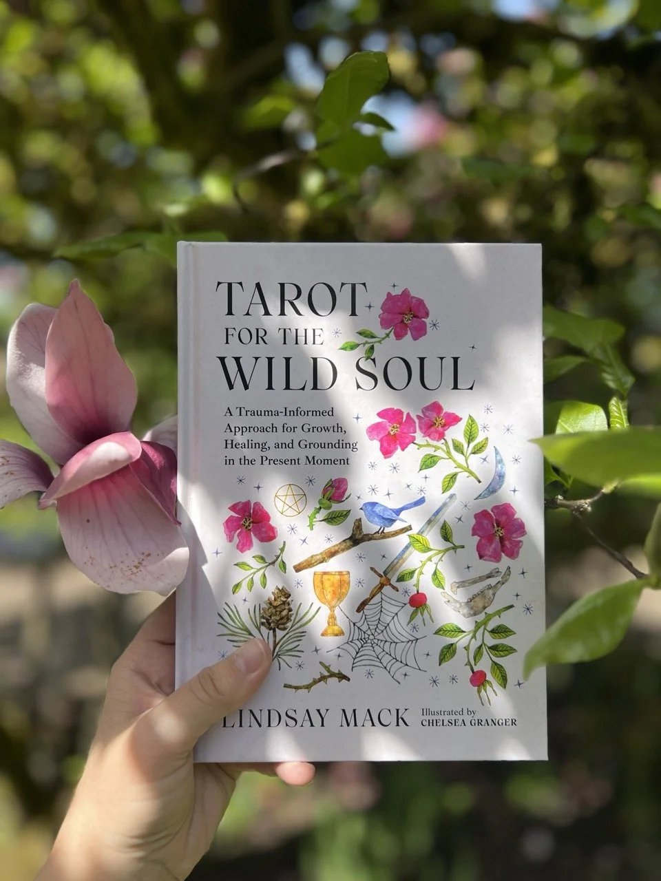 An image of someone's hand holding the book Tarot for the Wild Soul in front of green leaves and a pink flower.