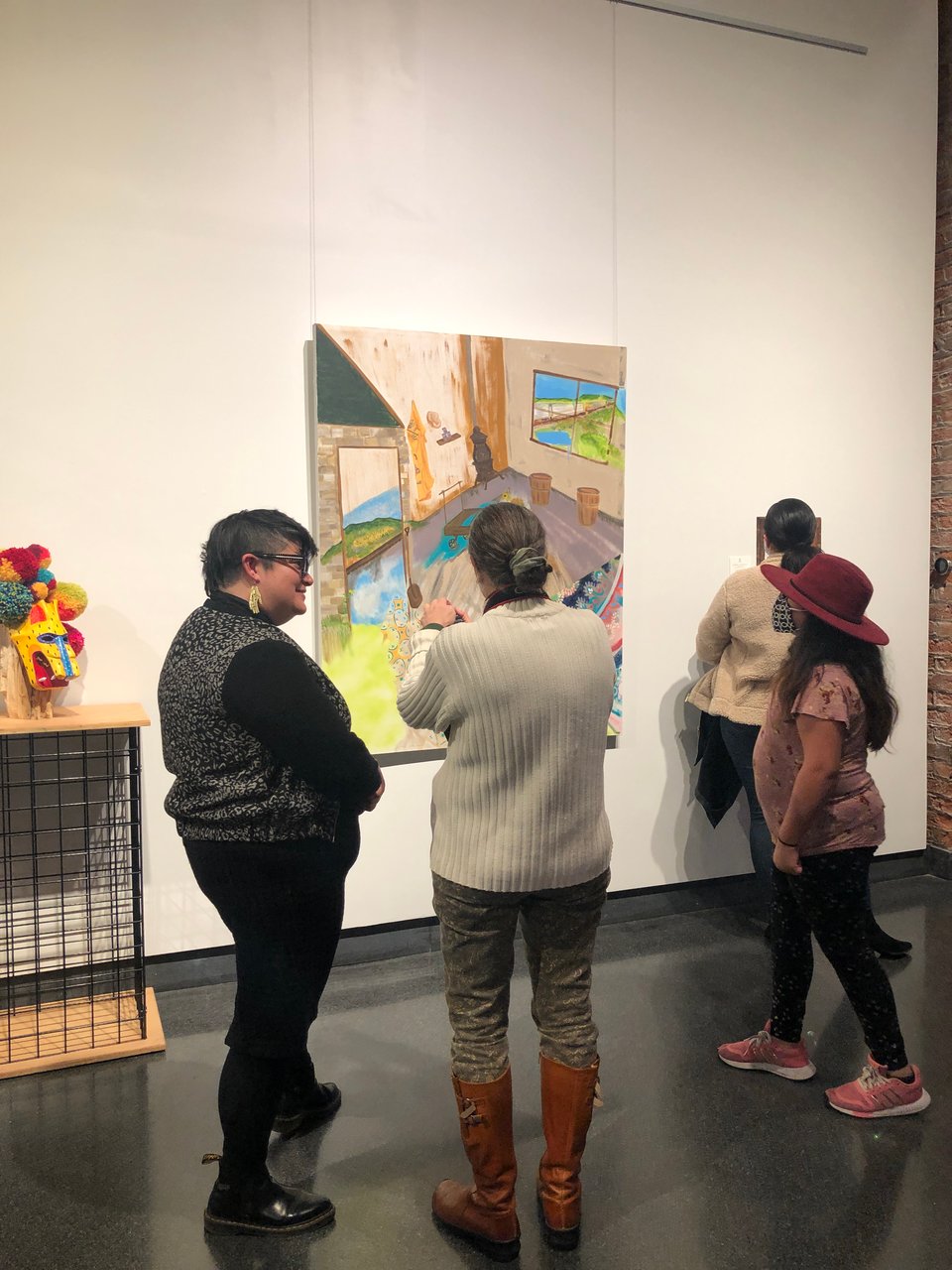 two femme people stand in front of an Art of KCF railroad themed painting discussing it, a sculpture is off to the side of the frame of a wooden animal mask with pompoms