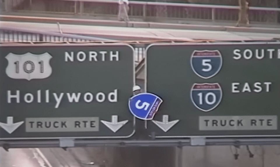 A man in a hard hat walking on a large freeway sign holding a sign reading Interstate 5 on it