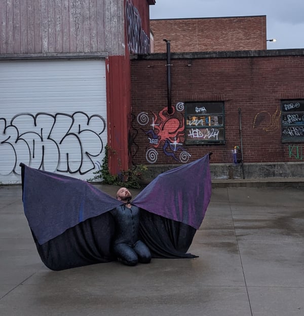I am kneeling on wet concrete in an industrial area, brick and loading bay and graffiti behind me. I have a huge glittery cape on, draping over my upstretched arms extended by my crutches. It is shimmery purple-pink-blue and frames me like wings. One of the graffiti items behind me is a swirling cosmic octopus.