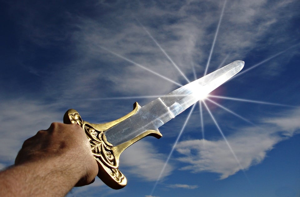 The left hand of a man holding a sword up to the sky; the sky is blue with a few cirrus clouds, and sun is glinting off the sword