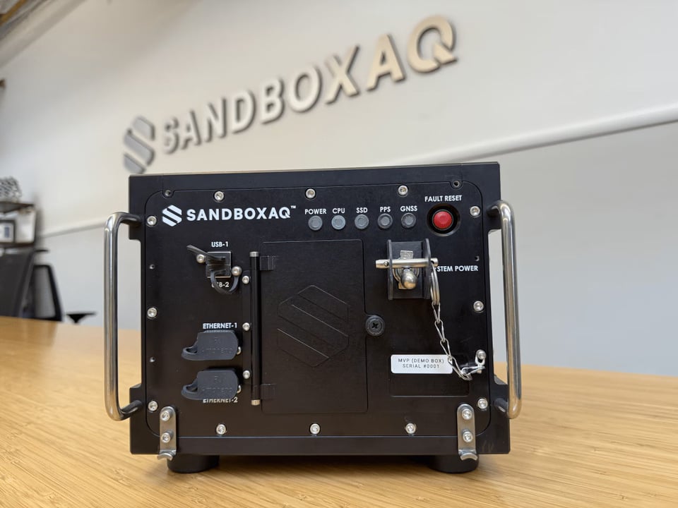 A black electronic device labeled “SandboxAQ” sits on a wooden table in an office with the same logo on the wall behind it. The device features metal handles, ports for USB and Ethernet connections, indicator lights, and a red fault reset button, giving it the appearance of ruggedized scientific or computing equipment.