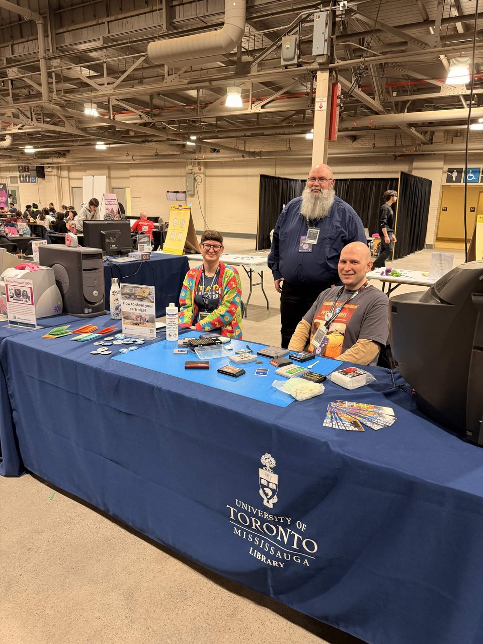 Three people at a University of Toronto Mississauga Library booth inside a large convention hall — a woman in a colourful patterned cardigan and a man in a grey T-shirt are seated behind a long table draped in a navy blue cloth bearing the university crest, while a bearded man in a blue shirt with a lanyard stands between them. The table displays retro computer equipment, game cartridges, cleaning supplies, pin badges and informational flyers, with vintage desktop towers and monitors on either end.
