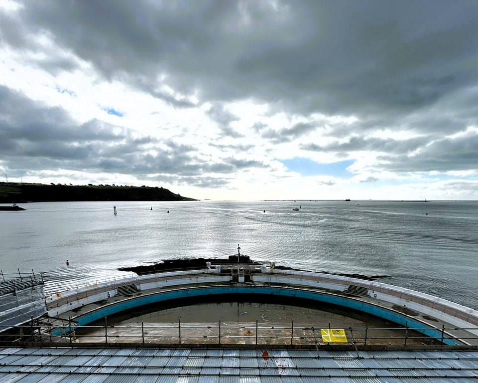 Looking across the Lido pool to Plymouth Sound on a grey day. The pool is half-filled with greenish water, and there are pools on the bath. In the foreground, tin roofed scaffolding hides the sun terrace.
