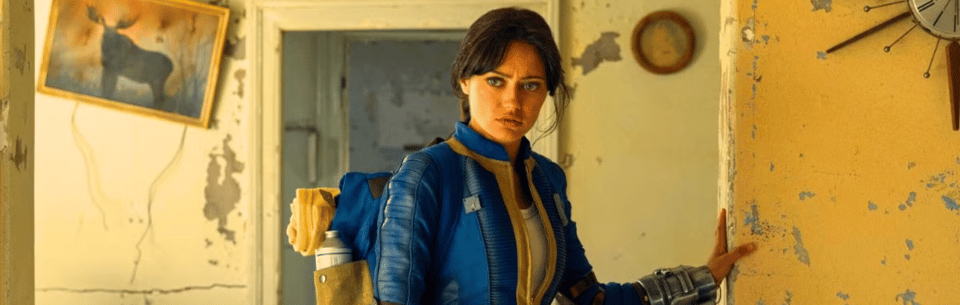 A still from Fallout, season 1. A white woman with brown hair stands in a sandblasted, decaying house. She wears a blue and yellow jumpsuit and looks quite concerned.
