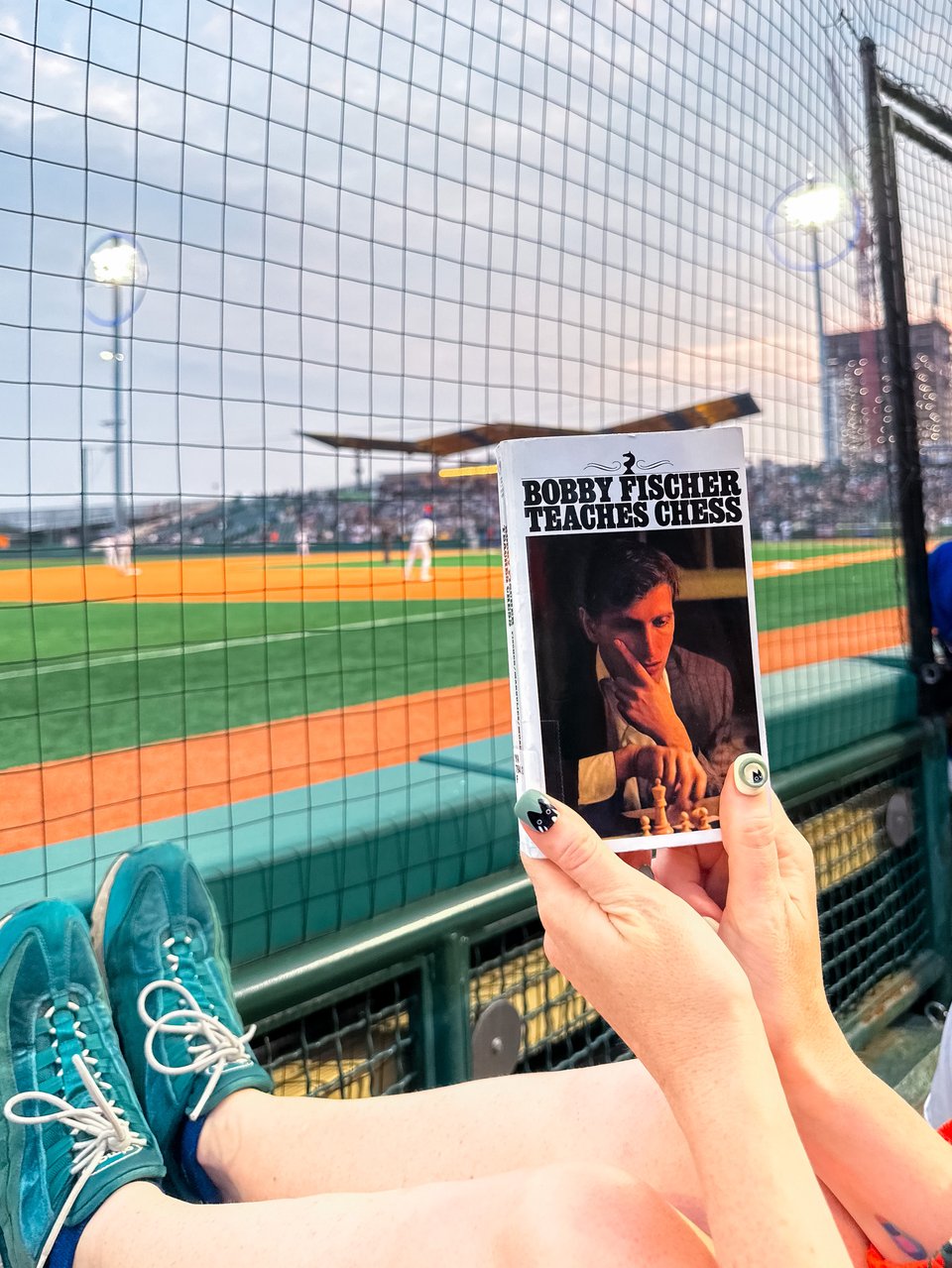 beth holds "Bobby Fischer teaches chess" up in front of the baseball game