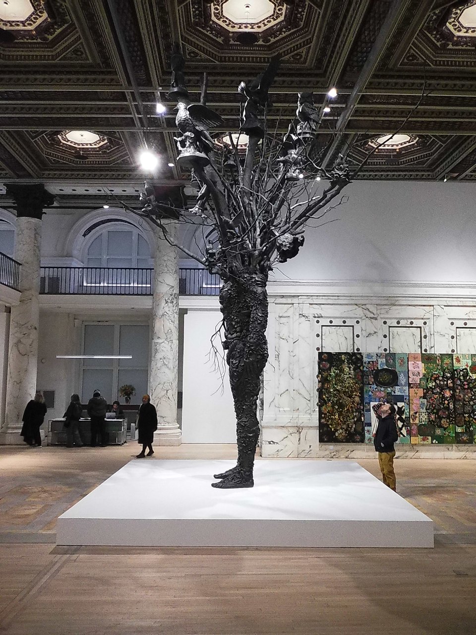 photo of gallery exhibition with a huge bronze cast of a man whose body morphs from the shoulders above into branches with flowers and birds on them. The statue is on a white square pedestal, and a man is standing next to it, looking up at the back of the figure